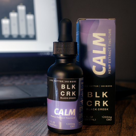 Black Creek CBD Calm Tincture standing with its box in front of a computer with work on it. Black background, wood surface